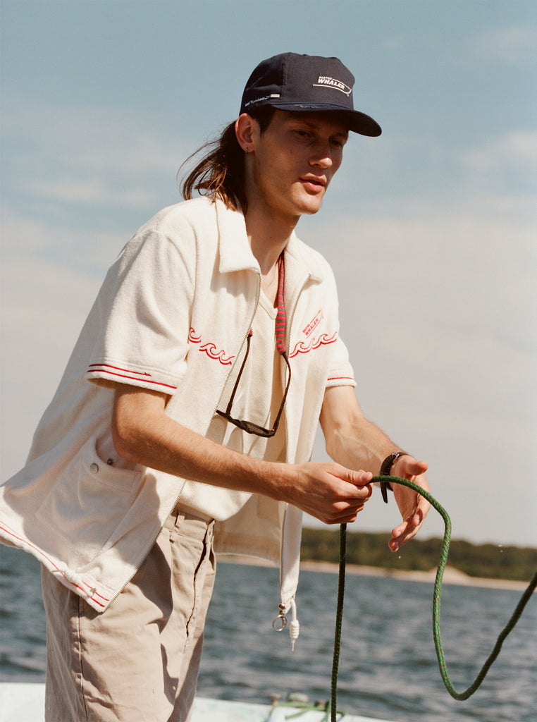 Male model with shoulder length brown hair wearing the 'Boston Whaler' 5-Panel Souvenir Cap while holding a line on a boat, shot on a background of the water, a thin strip of land, and blue skies.