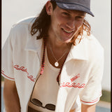Male model with shoulder length brown hair wearing the 'Boston Whaler' Cabana (white) unzipped with a white tank top underneath, and a navy boston whaler souvenir cap, shot on a background of the water, a strip of land, and blue skies.