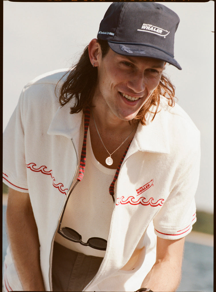 Male model with shoulder length brown hair wearing the 'Boston Whaler' Cabana (white) unzipped with a white tank top underneath, and a navy boston whaler souvenir cap, shot on a background of the water, a strip of land, and blue skies.