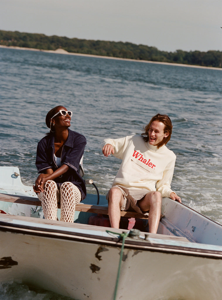 Male model sitting on the back of a boat wearing the 'Boston Whaler' Sweater while sitting next to a female model wearing a Boston Whaler cabana in navy.