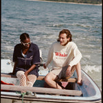 Female model and male model sitting together on a Boston Whaler, with the female model wearing the 'Boston Whaler' Cabana (navy), shot on a background of the water, a thin strip of land, and blue skies.