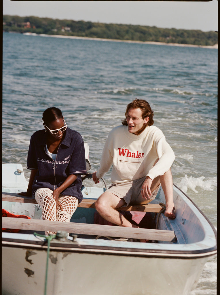 Female model and male model sitting together on a Boston Whaler, with the female model wearing the 'Boston Whaler' Cabana (navy), shot on a background of the water, a thin strip of land, and blue skies.