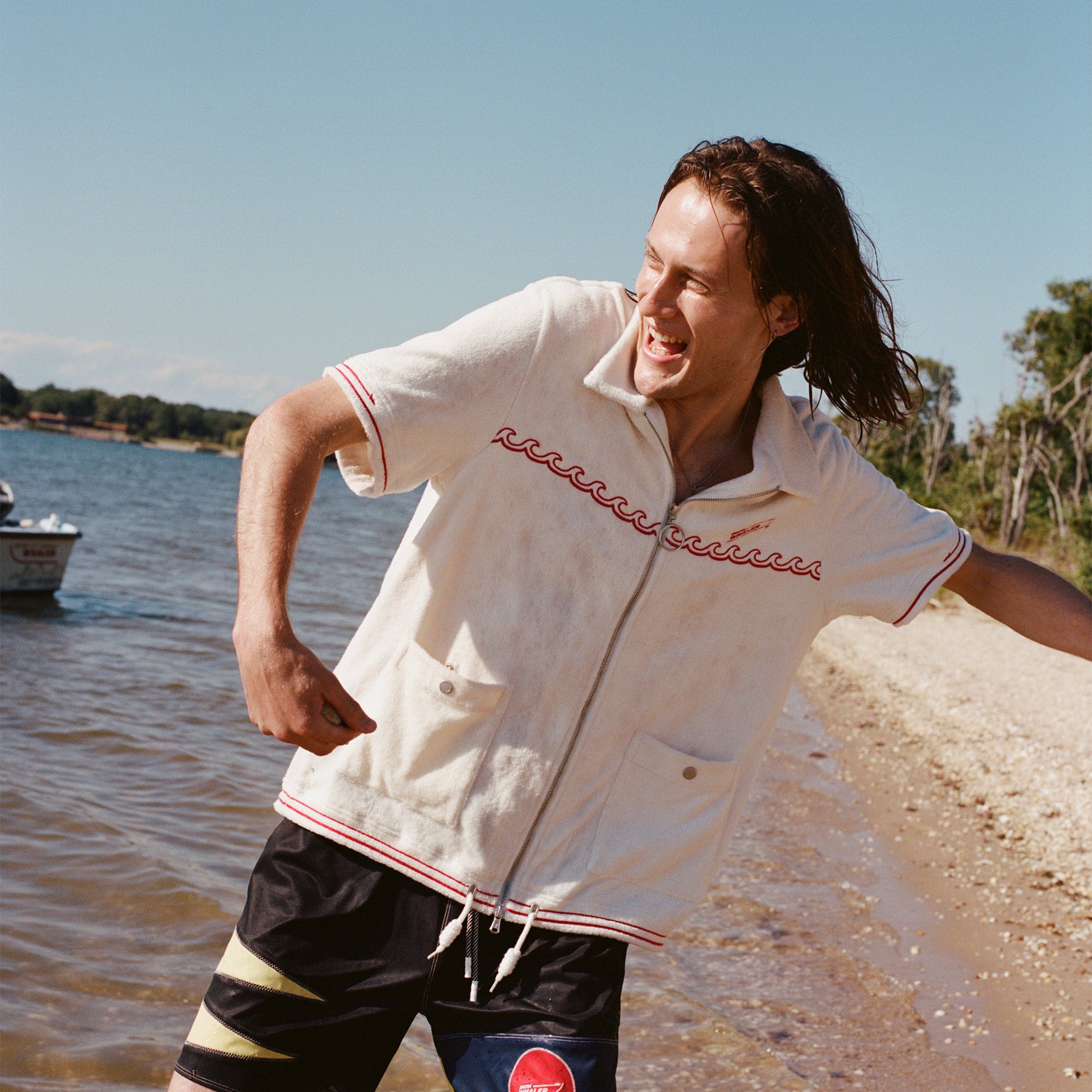 Male model wearing the 'Boston Whaler' swim trunks with a white boston whaler cabana as he skips rocks on the beach.