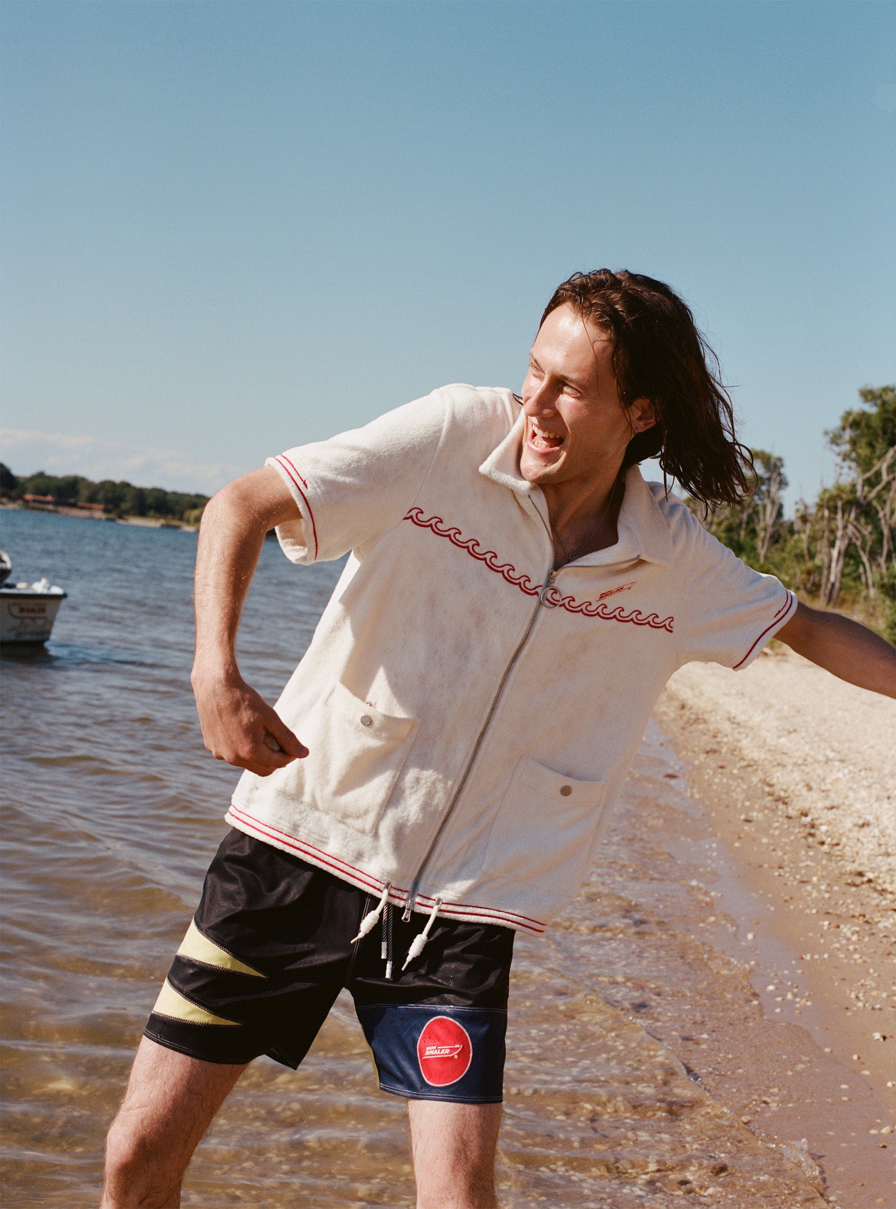 Male model wearing the 'Boston Whaler' swim trunks with a white boston whaler cabana as he skips rocks on the beach.