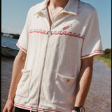 Male model with shoulder length brown hair wearing the 'Boston Whaler' Cabana (white) and black short, shot on a thin strip of land, and blue skies.