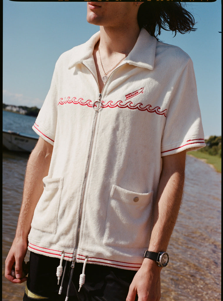 Male model with shoulder length brown hair wearing the 'Boston Whaler' Cabana (white) and black short, shot on a thin strip of land, and blue skies.