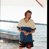 Male model with shoulder length brown hair wearing the 'boston whaler' no nibbles cabana with black swim trunks, shot on a background of water, a boston whaler anchored, a thin strip of land, and blue skies.