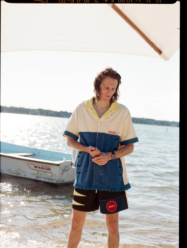 Male model with shoulder length brown hair wearing the 'boston whaler' no nibbles cabana with black swim trunks, shot on a background of water, a boston whaler anchored, a thin strip of land, and blue skies.