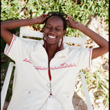 Female model seated wearing the 'Boston Whaler' Cabana (white) with white shorts and both arms holding onto sunglasses perched on the top of her head, shot on a background of greenery.