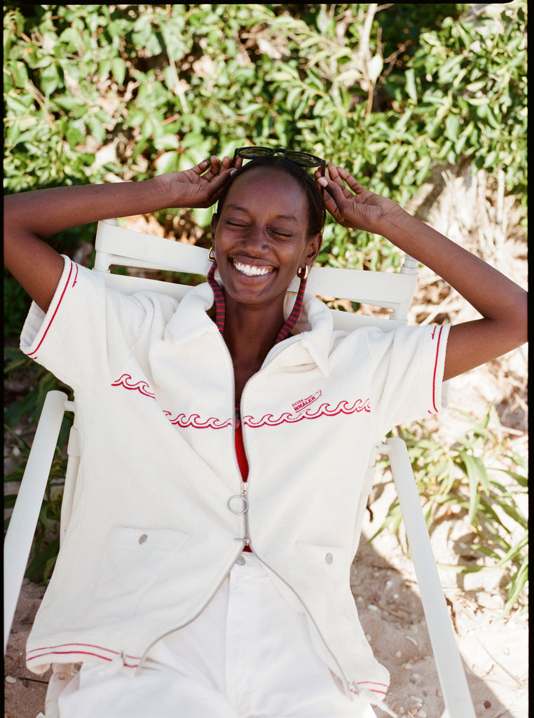 Female model seated wearing the 'Boston Whaler' Cabana (white) with white shorts and both arms holding onto sunglasses perched on the top of her head, shot on a background of greenery.