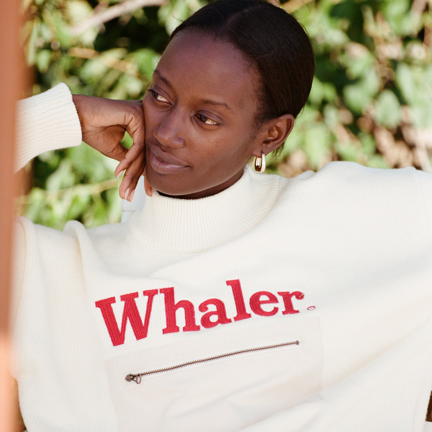 Female model with black hair wearing the 'Boston Whaler' Sweater, shot on a leafy green background.