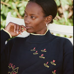 Person sitting in a white chair wearing a navy sweater with yellow and red fish designs and text. The sweater has yellow trim on the collar and cuffs. Green plants are in the background.