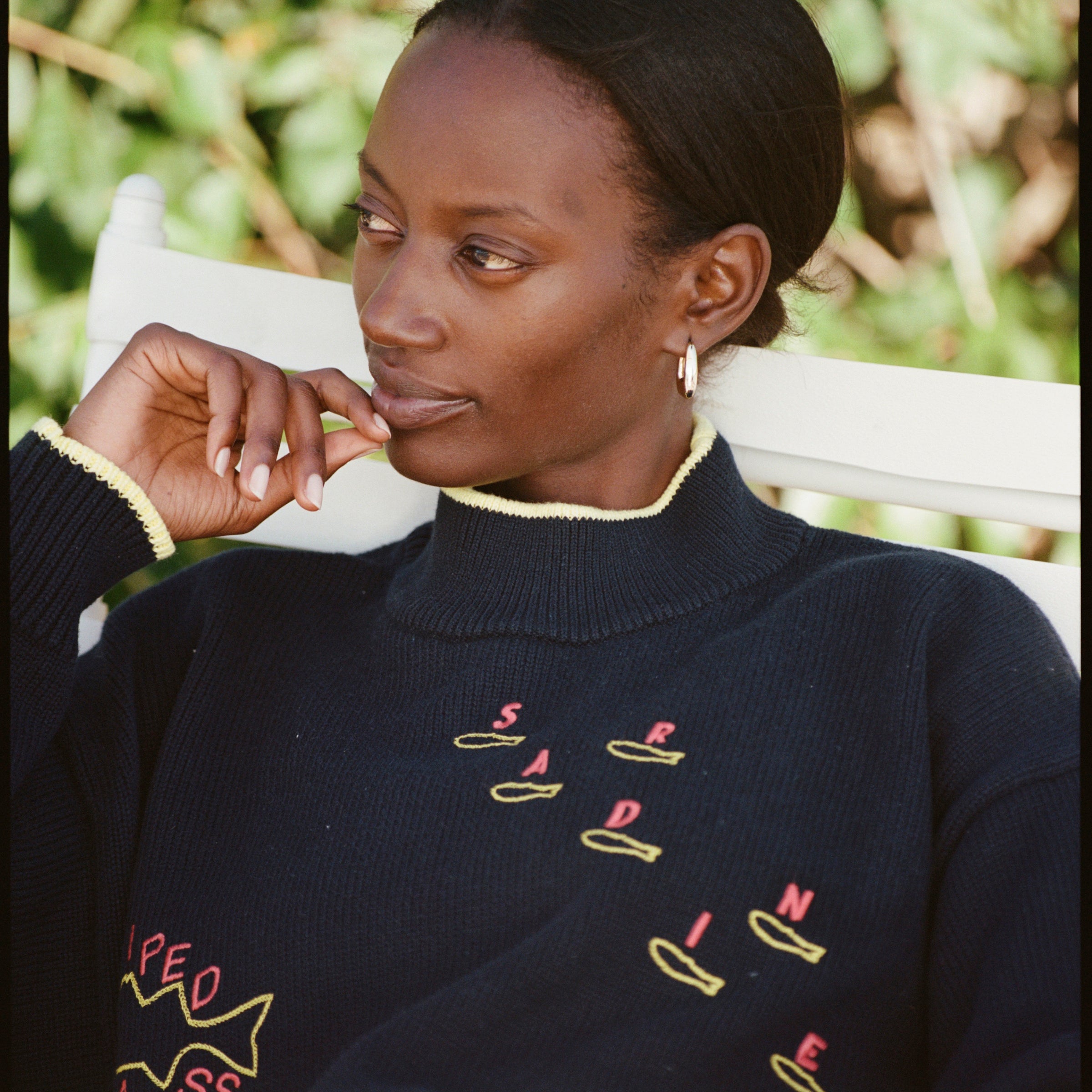 Person sitting in a white chair wearing a navy sweater with yellow and red fish designs and text. The sweater has yellow trim on the collar and cuffs. Green plants are in the background.