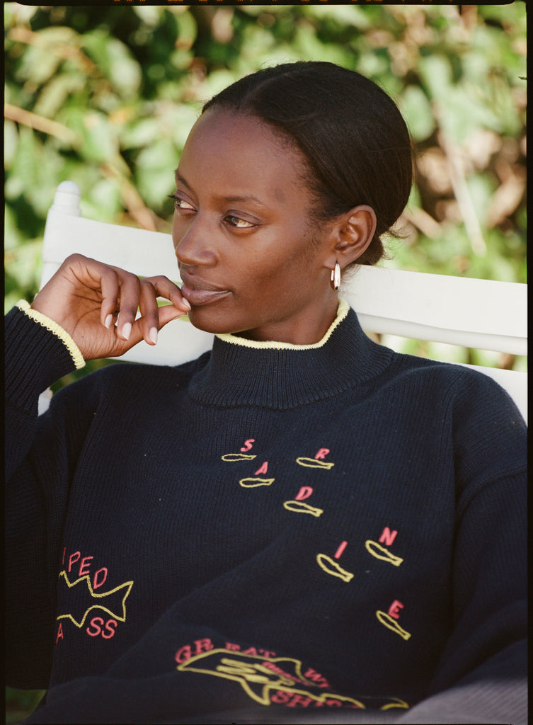 Person sitting in a white chair wearing a navy sweater with yellow and red fish designs and text. The sweater has yellow trim on the collar and cuffs. Green plants are in the background.