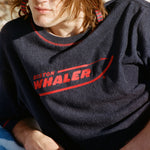 Male model with shoulder-length brown hair wearing the 'Boston Whaler' Souvenir Terry Tee while laying on the beach wearing sunglasses.