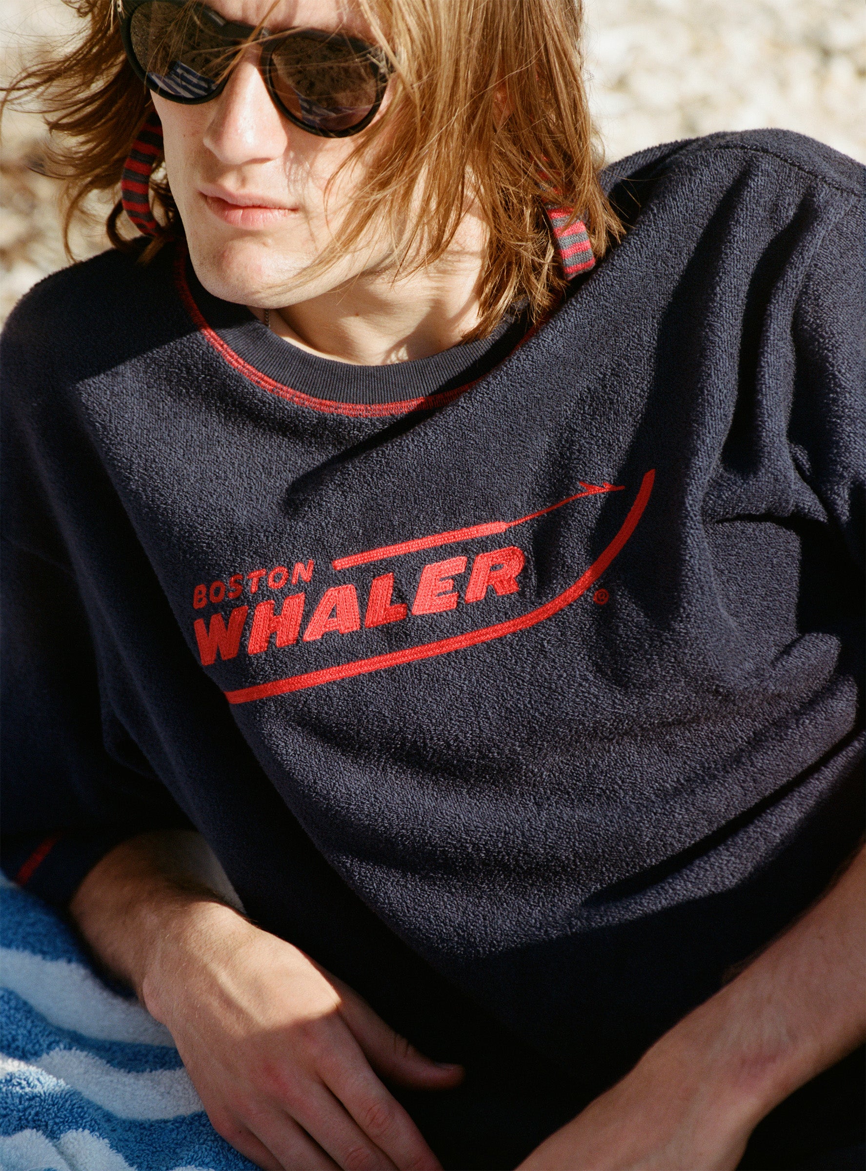 Male model with shoulder-length brown hair wearing the 'Boston Whaler' Souvenir Terry Tee while laying on the beach wearing sunglasses.