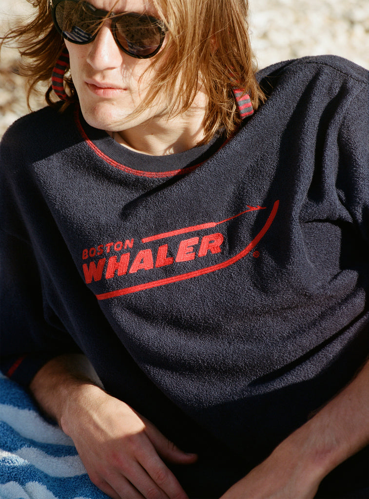 Male model with shoulder-length brown hair wearing the 'Boston Whaler' Souvenir Terry Tee while laying on the beach wearing sunglasses.