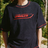 Male model with shoulder length brown hair wearing the 'Boston Whaler' Souvenir terry tee, shot on a backgroun dof beach and foliage.