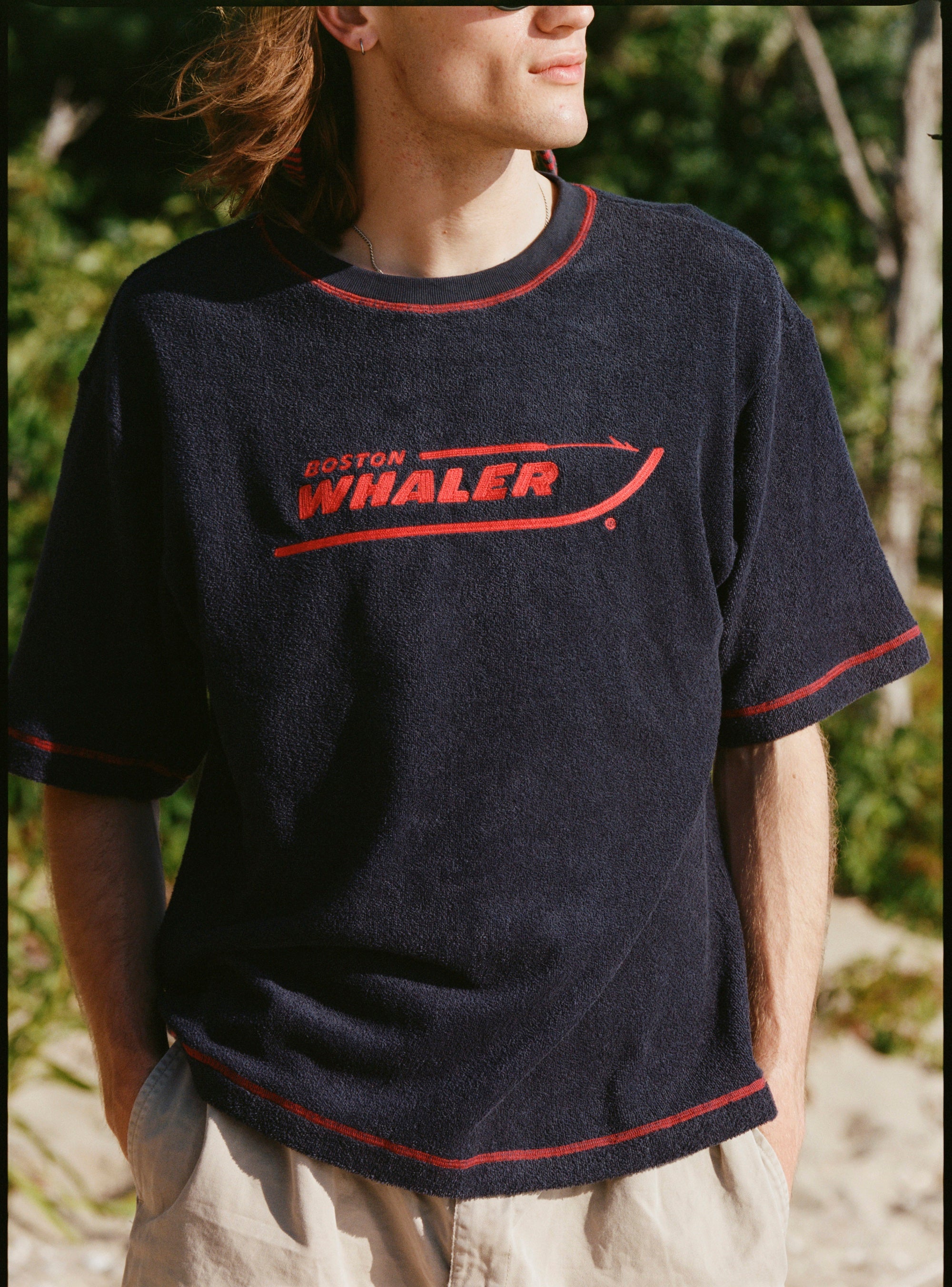 Male model with shoulder length brown hair wearing the 'Boston Whaler' Souvenir terry tee, shot on a backgroun dof beach and foliage.
