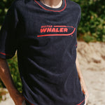 Male model with shoulder length brown hair turned to the side wearing the 'Boston Whaler' Souvenir Terry tee, shot on a background of the beach and foliage.