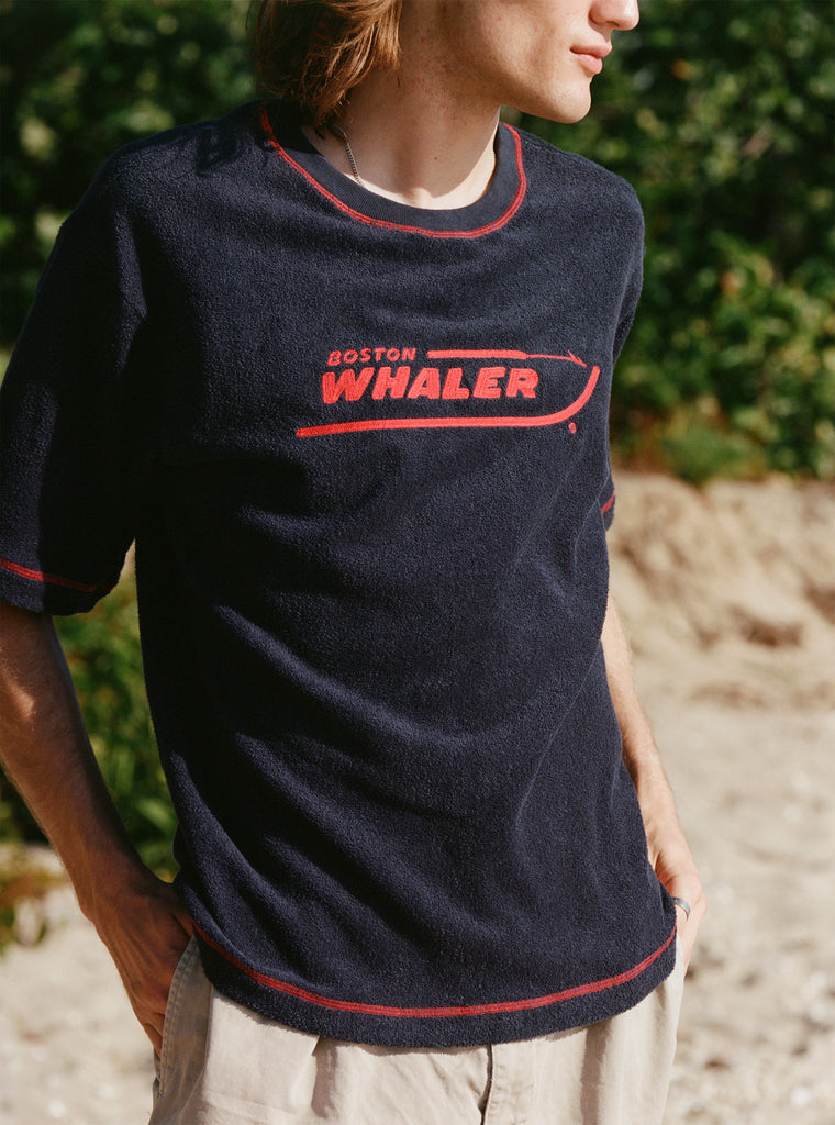 Male model with shoulder length brown hair turned to the side wearing the 'Boston Whaler' Souvenir Terry tee, shot on a background of the beach and foliage.