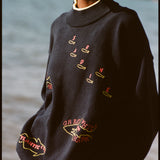 A woman wearing a navy knit sweater with yellow trim at the collar, cuffs, and hem. The sweater features playful, embroidered fish outlines with red lettering, including “Sardine” scattered across the chest, “Atlantic Cod” near the hem, and “Great White Shark (Human!)” on the lower torso. 