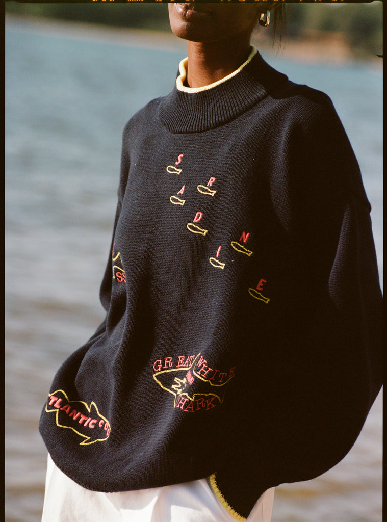 A woman wearing a navy knit sweater with yellow trim at the collar, cuffs, and hem. The sweater features playful, embroidered fish outlines with red lettering, including “Sardine” scattered across the chest, “Atlantic Cod” near the hem, and “Great White Shark (Human!)” on the lower torso. 