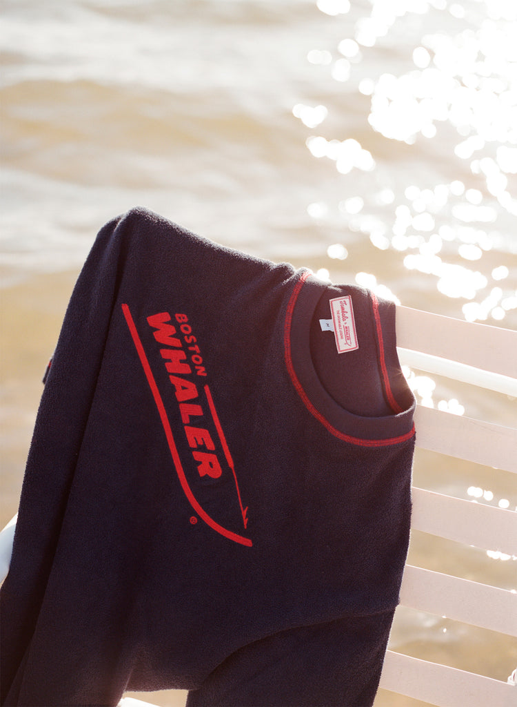 Picture of the 'boston whaler' souvenir terry tee hanging off of a white bench, shot on a background of glistening water.