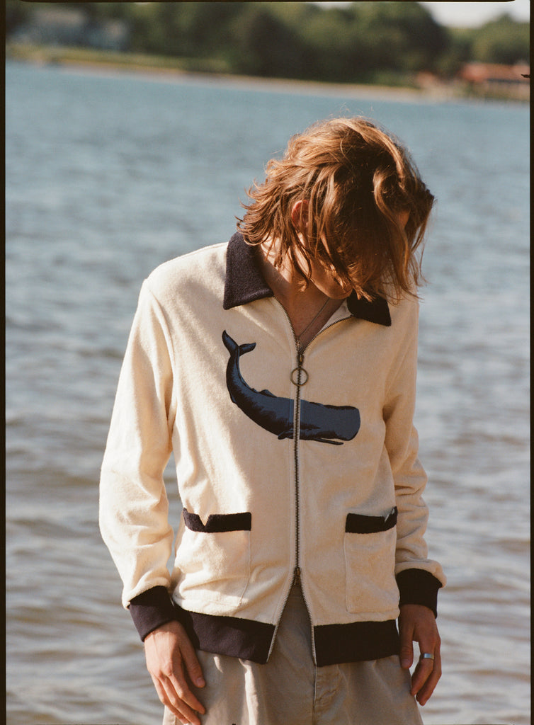 Man with long blonde hair looking down in front of water, wearing the long sleeve 'Blubber' Cabana.