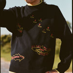 A man wearing a navy knit sweater with yellow trim at the collar, cuffs, and hem. The sweater features playful, embroidered fish outlines with red lettering, including “Sardine” scattered across the chest, “Atlantic Cod” near the hem, and “Great White Shark (Human!)” on the lower torso. 