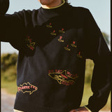 A man wearing a navy knit sweater with yellow trim at the collar, cuffs, and hem. The sweater features playful, embroidered fish outlines with red lettering, including “Sardine” scattered across the chest, “Atlantic Cod” near the hem, and “Great White Shark (Human!)” on the lower torso. 