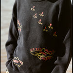 A person wearing a navy knit sweater with yellow trim at the collar, cuffs, and hem. The sweater features playful, embroidered fish outlines with red lettering, including “Sardine” scattered across the chest, “Atlantic Cod” near the hem, and “Great White Shark (Human!)” on the lower torso. 
