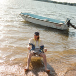 Male model seated in the water wearing a 'Boston Whaler' No nibbles cabana unbuttoned with a white shirt underneath and khaki shorts, shot on a background of water, a boston whaler anchored, a thin strip of land, and blue skies.