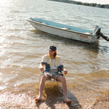 Male model seated in the water wearing a 'Boston Whaler' No nibbles cabana unbuttoned with a white shirt underneath and khaki shorts, shot on a background of water, a boston whaler anchored, a thin strip of land, and blue skies.