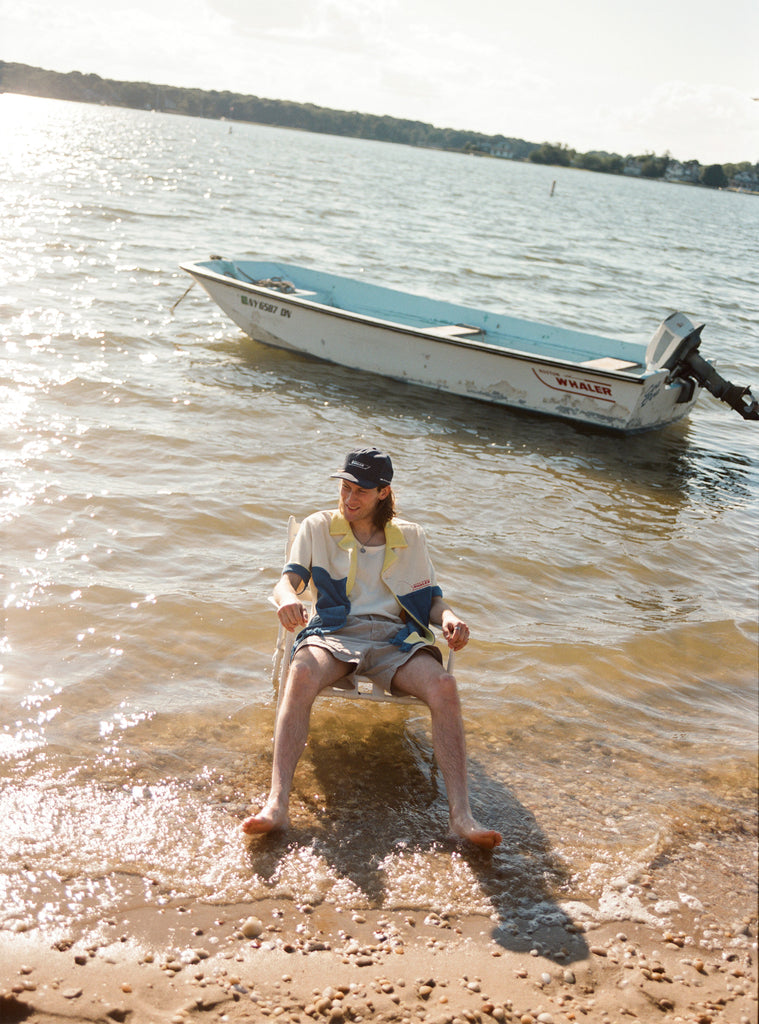 Male model seated in the water wearing a 'Boston Whaler' No nibbles cabana unbuttoned with a white shirt underneath and khaki shorts, shot on a background of water, a boston whaler anchored, a thin strip of land, and blue skies.