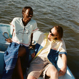 Female model with brown hair wearing the 'Boston Whaler' Hand Towel Top buttoned with a navy dress alongside a male model with brown shoulder-length hair, shot on a boat on the water.