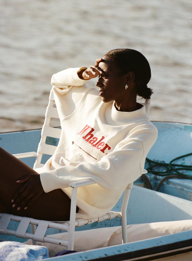 Female model seated wearing the 'Boston Whaler' sweater using her hand to shade her eyes from the sun on a boat, shot on the water.