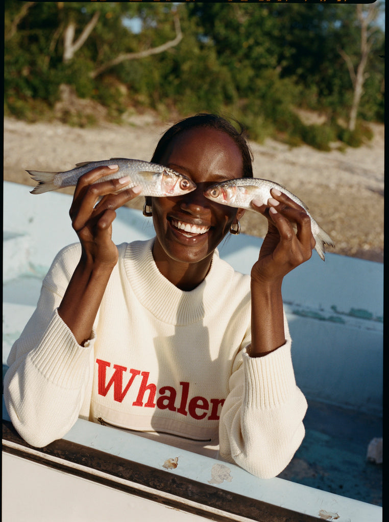 Female model holding two small fish to her face so that their eyes are in the position hers would be wears the 'Boston Whaler' Sweater while on a boat docked by a beach with foliage.