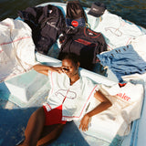 Female model lying on a boat while wearing the 'Boston Whaler' Hand Towel Top buttoned with a red swimsuit underneath while she is surrounded by various pieces from the Boston Whaler Tombolo collaboration.