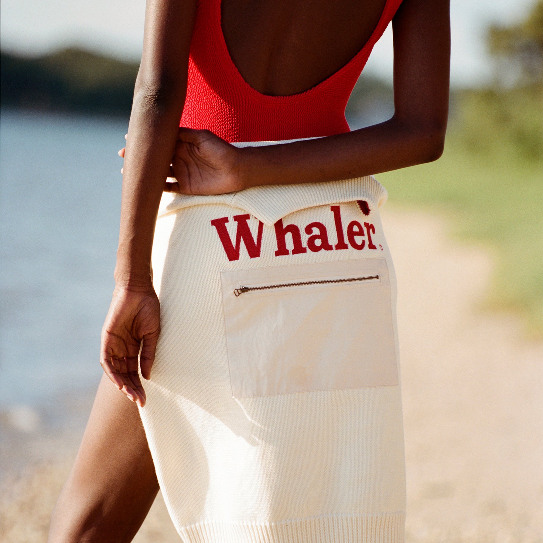 Female model facing away from the camera with a 'Boston Whaler' sweater tied around her waist so that the front faces toward the camera, wearing a red swimsuit while standing on the beach.