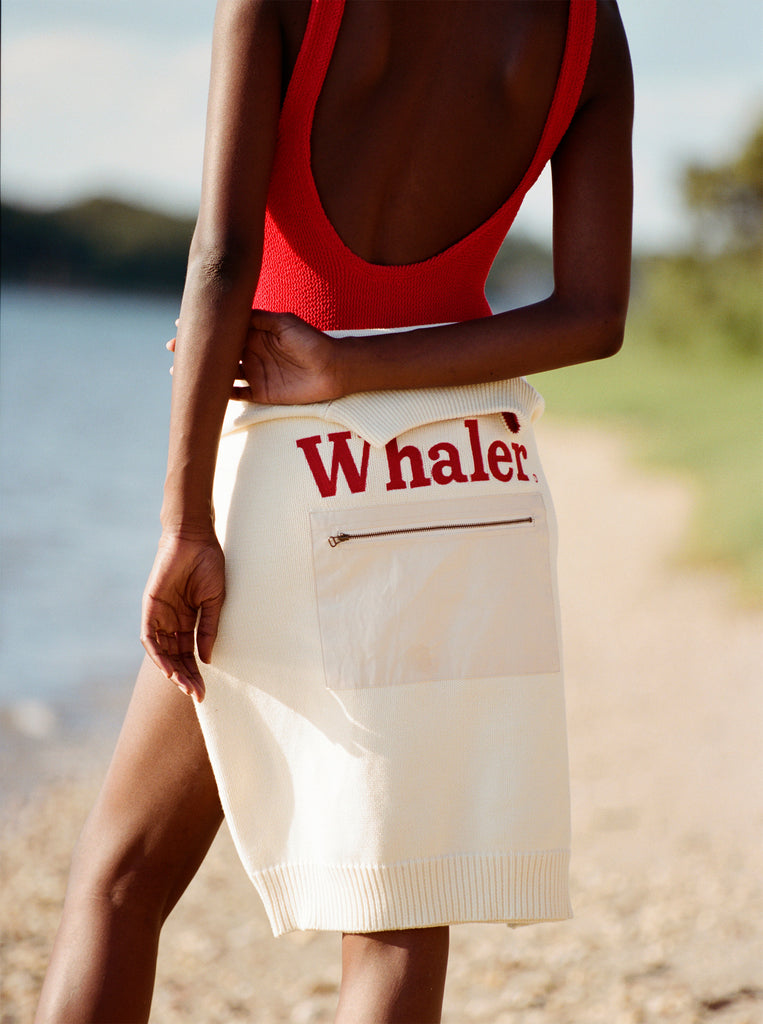 Female model facing away from the camera with a 'Boston Whaler' sweater tied around her waist so that the front faces toward the camera, wearing a red swimsuit while standing on the beach.
