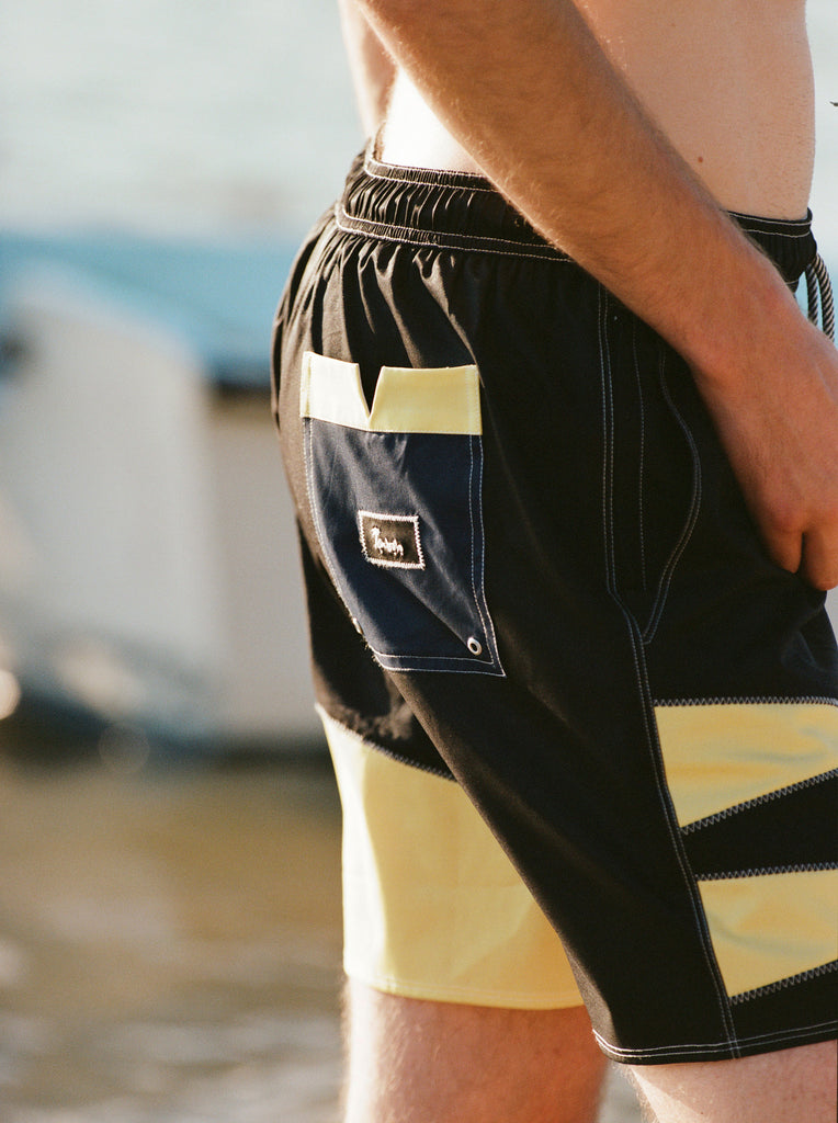 Male model wearing the 'Boston Whaler' Swim trunks while facing away from the camera to showcase the Tombolo patch on the back pocket that also has the signature Tombolo V-slit.