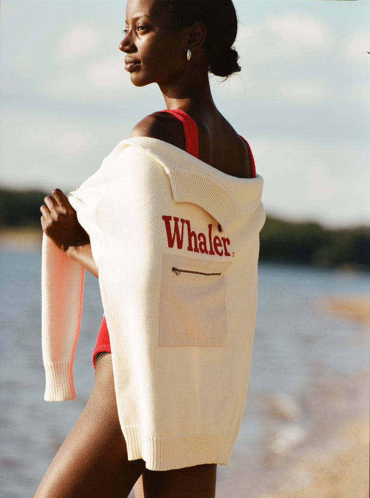 Female model with black hair in a bun stands facing away from the camera while holding her 'Boston Whaler' sweater over her back by the sleeves, wearing a red swimsuit, shot against a beach and blue skies.