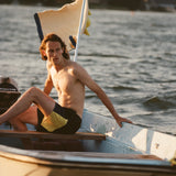 Male model on a boat wearing the 'boston whaler' swim trunks, shot on a background of the water, a flag fluttering in the wind, a thin strip of land, and blue skies.