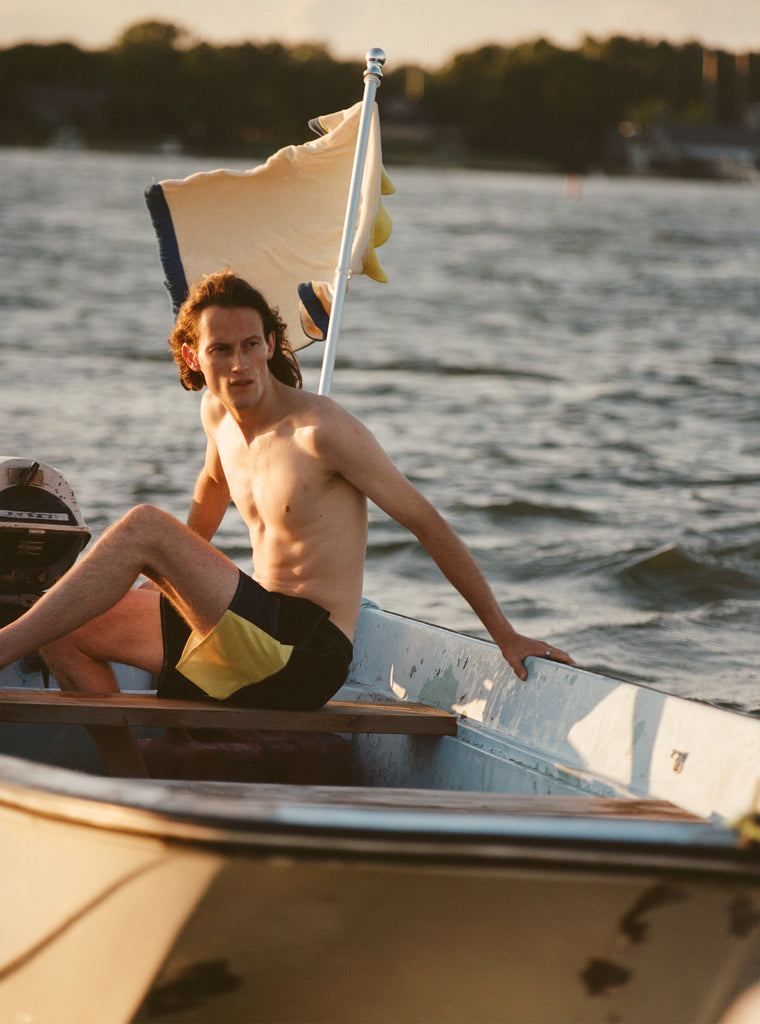 Male model on a boat wearing the 'boston whaler' swim trunks, shot on a background of the water, a flag fluttering in the wind, a thin strip of land, and blue skies.