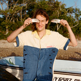 Male model holding to fish up to his face while wearing the 'Boston Whaler' No Nibbles Cabana, standing in front of boat resting on a beach with trees and blue skies.