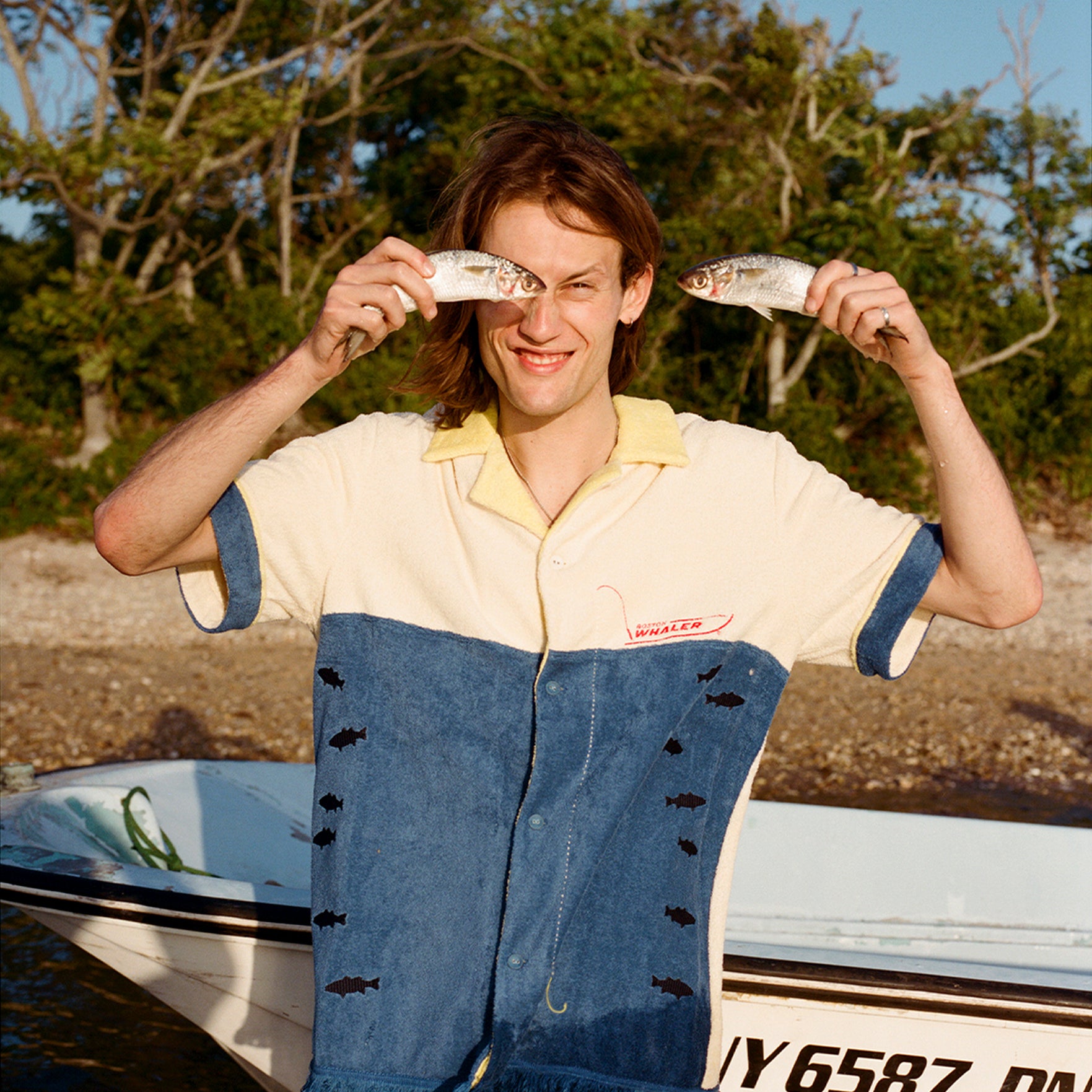 Male model holding to fish up to his face while wearing the 'Boston Whaler' No Nibbles Cabana, standing in front of boat resting on a beach with trees and blue skies.