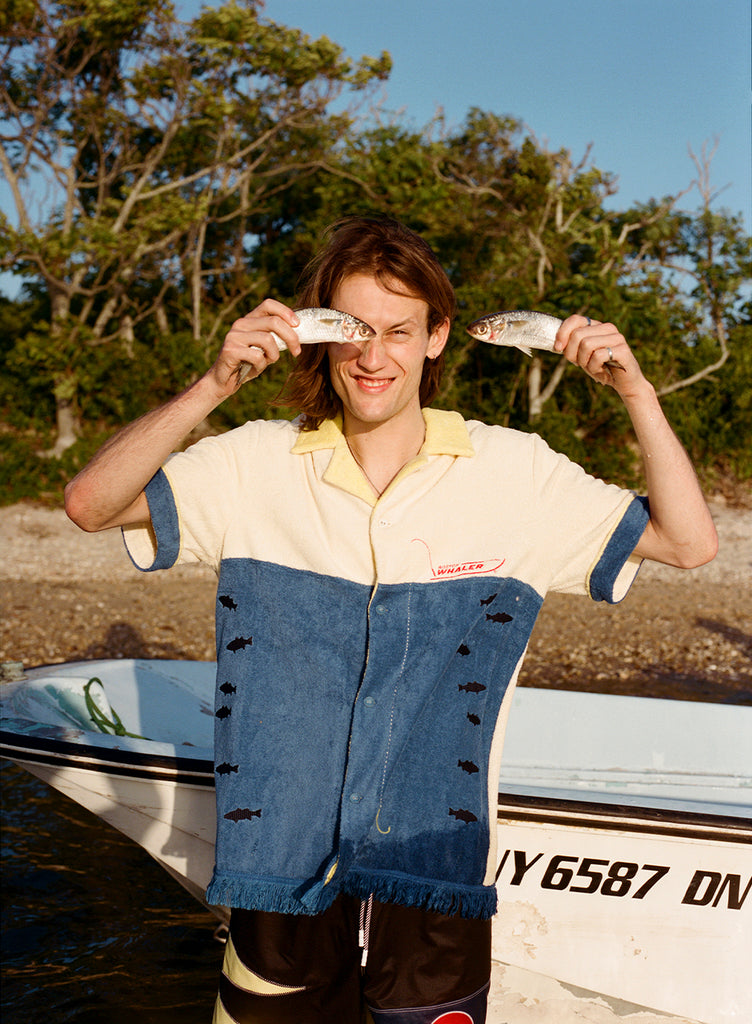 Male model holding to fish up to his face while wearing the 'Boston Whaler' No Nibbles Cabana, standing in front of boat resting on a beach with trees and blue skies.