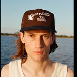 Man wearing Tombolo brown cap with a navy blue brim. The front features white embroidered text that reads “TOMBOLO” with a seashell illustration forming the letter “T,” and below it, “732 MONTAUK HIGHWAY.”