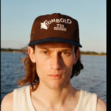 Man wearing Tombolo brown cap with a navy blue brim. The front features white embroidered text that reads “TOMBOLO” with a seashell illustration forming the letter “T,” and below it, “732 MONTAUK HIGHWAY.”